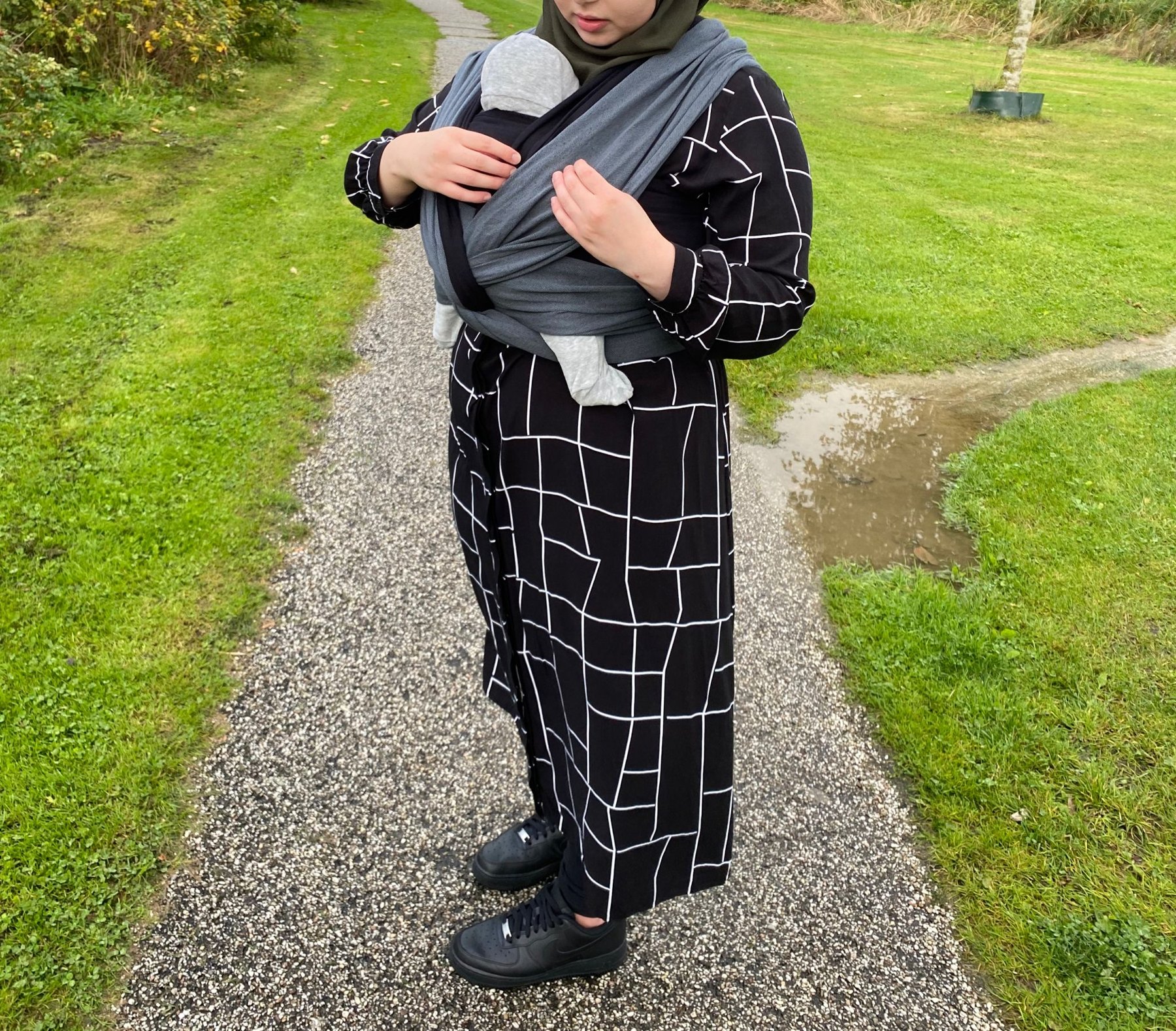 Baby wordt op de buik gedragen in een grijze geweven draagdoek op een pad