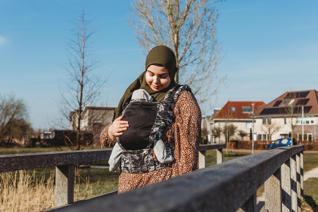 Feride kijkt naar haar baby die zij in een draagzak draagt, buiten bij een hek