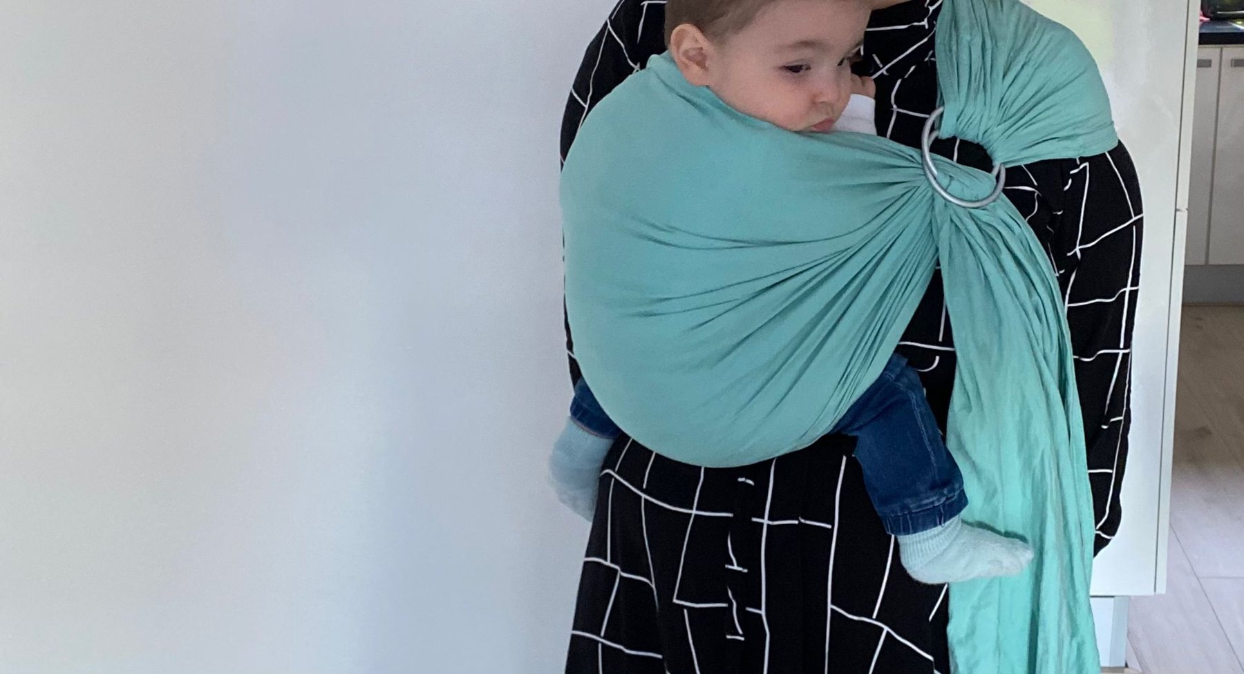 Kind wordt binnenshuis gedragen in een turquoise ringsling en kijkt opzij